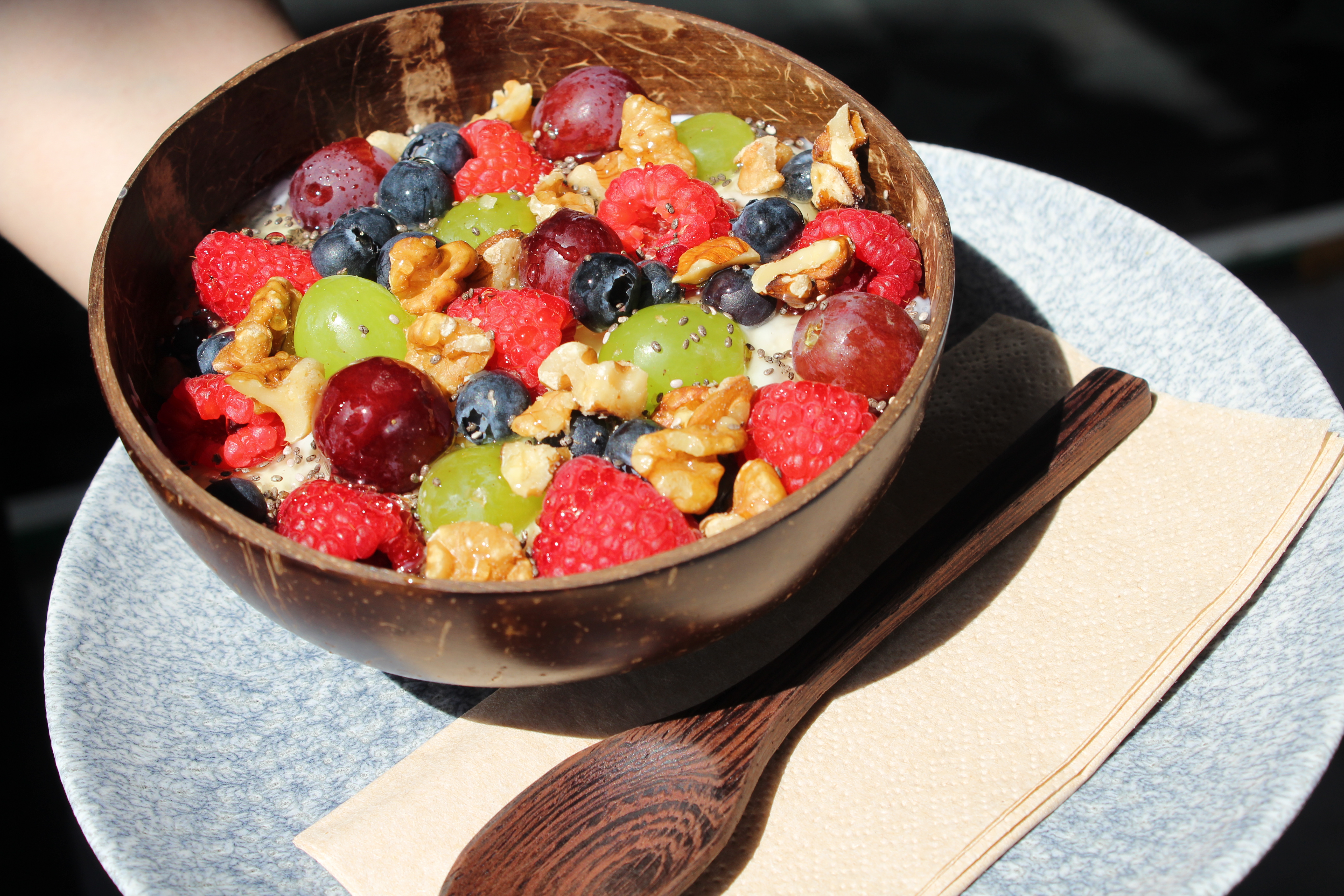 A bowl filled with assorted fresh fruits, including raspberries, blueberries, green grapes, and walnuts, placed on a blue plate with a wooden spoon and napkin.